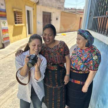 Das Bild zeigt eine Frau, die ein Foto mit zwei Frauen in traditioneller Kleidung macht, die auf einer von Gebäuden gesäumten Straße stehen, wahrscheinlich während einer kulturellen oder kommunalen Veranstaltung.