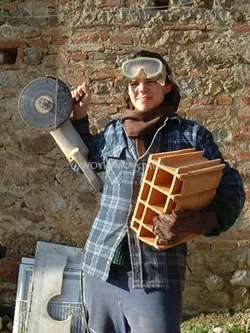 The image depicts a man standing in front of a brick wall, holding a circular saw and a stack of bricks, wearing a plaid shirt, goggles, and gloves.