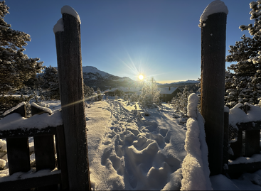 A snowy landscape with deep footprints leads through a gate toward sunlit cabins and mountains, surrounded by trees, under a clear blue winter sky at sunrise.