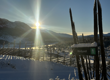 A bright winter sunrise over snowy mountains and a wooden fence. A "Hindjuvet" hiking trail sign points ahead, creating a peaceful, scenic outdoor atmosphere.