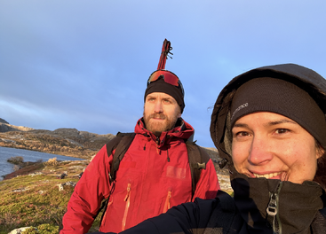 A man and woman in outdoor winter gear smile for a selfie. Behind them, a scenic landscape with hills, water, and blue sky bathed in sunlight.