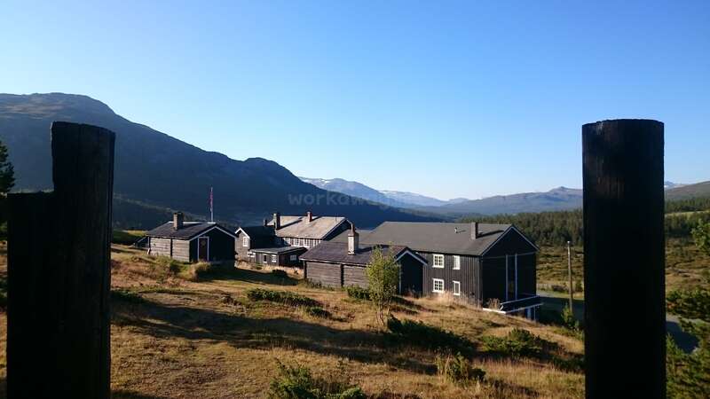 The image depicts a serene rural landscape featuring a cluster of houses with dark roofs and chimneys, set against a backdrop of majestic mountains under a clear blue sky.