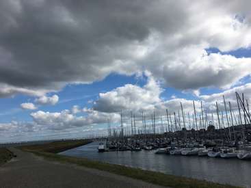 L'image représente une scène de marina sereine, avec une rangée de bateaux amarrés au bord de l'eau, sur fond de ciel nuageux et de berge herbeuse.