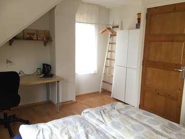 L'image représente une chambre à coucher compacte avec un bureau, un lit et une armoire, avec des murs blancs, du parquet et une fenêtre avec un rideau blanc.