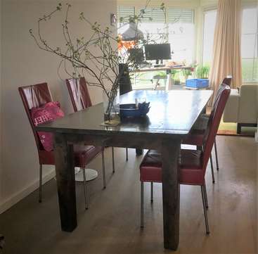 L'image représente une salle à manger avec une table en bois foncé et quatre chaises rouges, avec un vase de branches et un bureau d'ordinateur à l'arrière-plan.