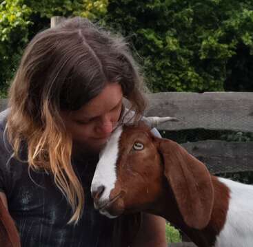 Eine Frau lehnt ihre Stirn liebevoll an eine braun-weiße Ziege und teilt einen zärtlichen Moment im Freien. Ein Holzzaun und grünes Laub bilden einen friedlichen Hintergrund.