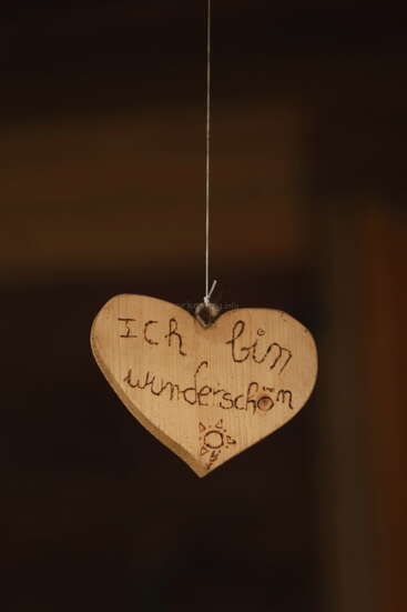 Ein herzförmiges Ornament aus Holz hängt an einer Schnur. Darauf sind die deutschen Worte "Ich bin wunderschön" und ein kleines Sonnensymbol eingraviert.