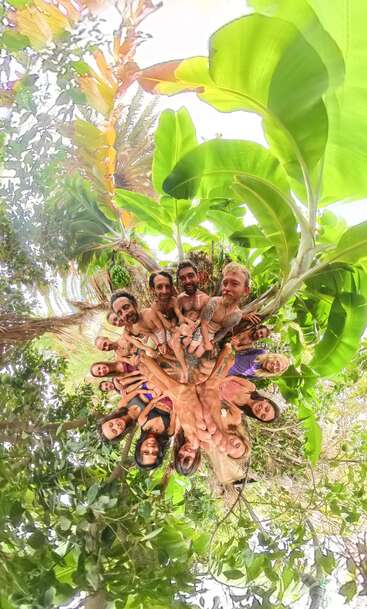 A joyful group of people stand close together in a lush, tropical setting, captured in a circular, panoramic view surrounded by vibrant green leaves above.
