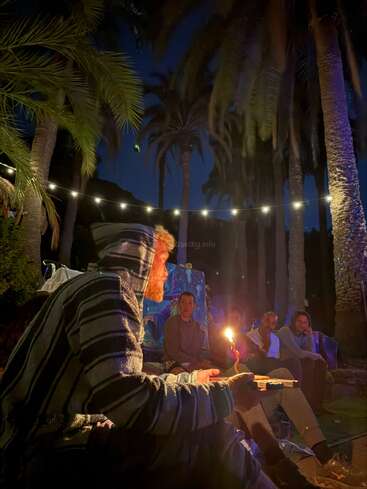 A group of people sit outdoors under palm trees at night, illuminated by string lights and a candle. One person in a striped hoodie is foregrounded.