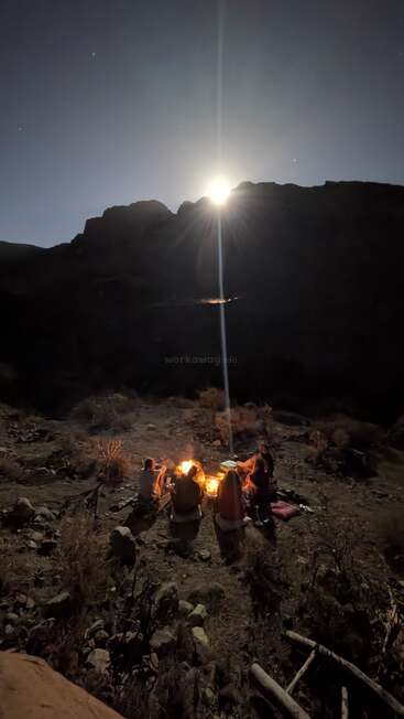 A group sits around a glowing campfire under the night sky, surrounded by desert landscape and mountains, illuminated by the rising moon beyond the dark silhouette.