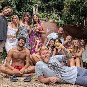 Eine Gruppe von dreizehn lächelnden Menschen posiert im Freien unter Bäumen, einige sitzend, einige stehend. Sie sehen glücklich und entspannt aus und genießen ein zwangloses Beisammensein, bei dem sie gemeinsam Obst essen.