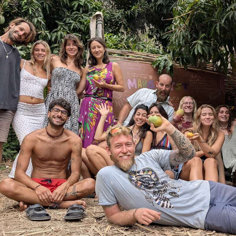 A group of thirteen smiling people pose outdoors under trees, some sitting, some standing. They appear happy, relaxed, and enjoy a casual gathering, holding fruits together.