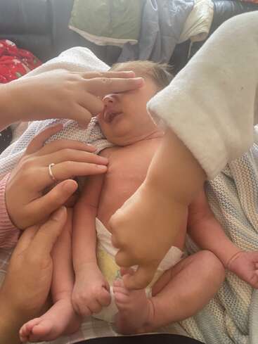 Un recién nacido tumbado en una manta, rodeado de manos cariñosas que lo tocan y lo sostienen suavemente, con una mano tapándole los ojos juguetonamente. Un momento acogedor.