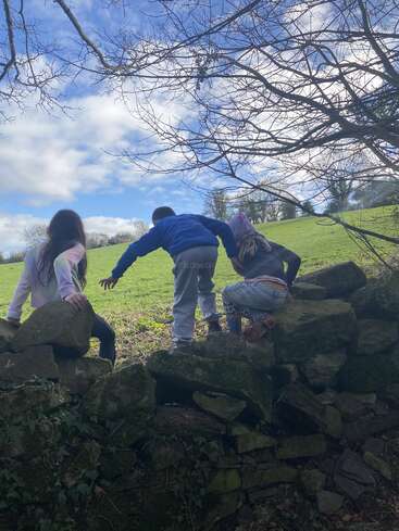 Tres niños trepan por un muro de piedra musgosa en dirección a un campo verde bajo un cielo parcialmente nublado con ramas desnudas en lo alto, lo que sugiere una aventura de principios de primavera.