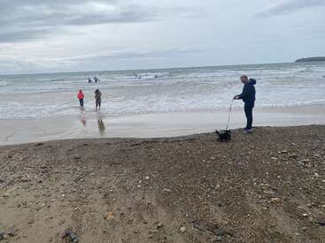 Un día nublado en la playa con gente cerca del agua, una persona paseando a un perro pequeño por la arena y olas al fondo.