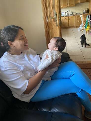 Una mujer sonriente se sienta en un sofá con un bebé en brazos, manteniendo el contacto visual. En la cocina, detrás de ellos, un pequeño perro negro mira hacia otro lado, sentado tranquilamente.