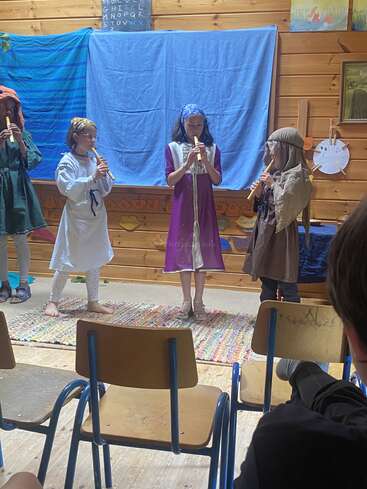 Cuatro niños disfrazados tocan la flauta dulce en el escenario durante una representación escolar. El público observa desde las sillas. El telón de fondo es azul con letras y dibujos.