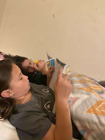 Dos niños están tumbados en la cama, leyendo juntos un libro. Uno sostiene una linterna, el otro está concentrado en las páginas, bajo una manta estampada.