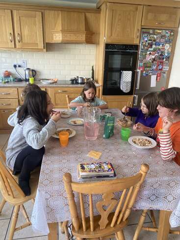 Cuatro niños se sientan alrededor de una mesa de cocina a comer. La cocina es acogedora, con armarios de madera, una nevera cubierta de imanes y libros sobre la mesa.
