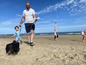 Un hombre pasea con un niño pequeño y un perro por una playa de arena. Los niños juegan y dan volteretas junto al mar bajo un cielo azul.