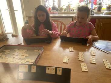 Dos chicas están sentadas a una mesa de madera, jugando a un juego de fichas. La luz del sol entra a raudales por la ventana de la cocina, iluminando el ambiente amistoso.