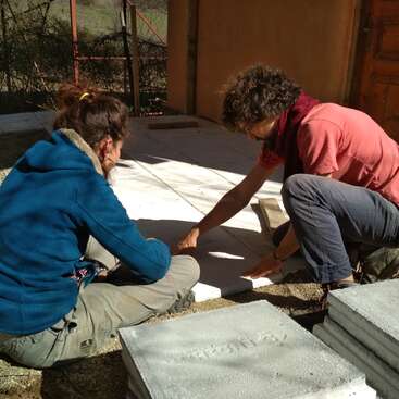 Deux personnes sont accroupies sur le sol, travaillant avec des blocs de béton devant un bâtiment en bois par une journée ensoleillée.