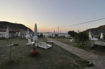 The image depicts a serene outdoor setting with a grassy area, a stone path, and a few white lounge chairs and umbrellas scattered about, set against a picturesque sunset backdrop.