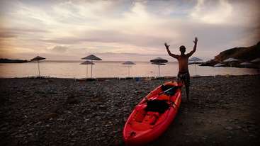 A person is standing on the shore of a body of water, with a kayak or canoe in the background. The image appears to be taken from a beach or a lake.
