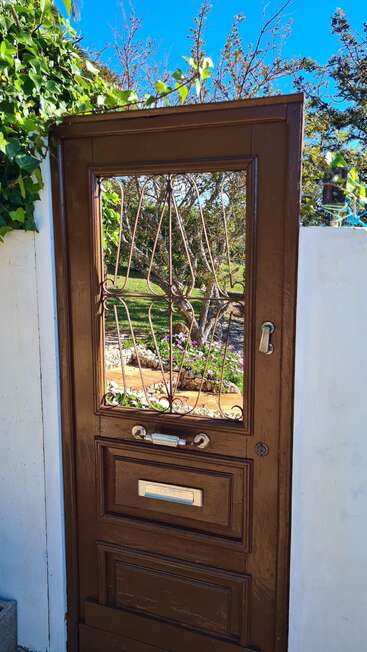 Una puerta de madera marrón con barrotes metálicos se alza en un muro blanco, parcialmente cubierto por enredaderas verdes. A través de ella se ve un colorido jardín y un árbol.