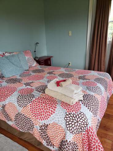 A cozy bedroom with pastel green walls, floral-patterned bedspread, pillows, neatly stacked towels, a bedside lamp, red nightstand, wooden floor, and brown curtains.
