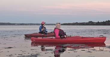 Two women are kayaking on a calm lake during sunset, wearing life jackets. The sky is pink and purple, reflecting beautifully on the tranquil water surface.