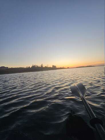 A calm lake at sunset features gentle rippling water, a kayak paddle in the foreground, and silhouetted trees and distant shoreline under a clear, expansive sky.