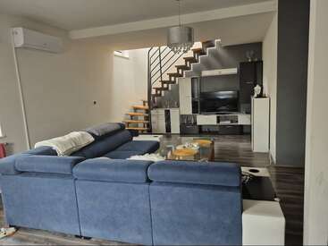 Ein modernes Wohnzimmer mit einem großen blauen Ecksofa, einem Couchtisch aus Glas, einem TV-Möbel, einer offenen Treppe, Holzböden, neutralen Wänden und eleganter Beleuchtung. Gemütliche Atmosphäre.