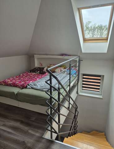 Ein gemütliches Schlafzimmer im Dachgeschoss verfügt über ein Bett mit bunter Bettwäsche, Plüschtiere, ein Dachfenster, ein kleines Regal und eine moderne Treppe mit Metallgeländer, die nach unten führt.