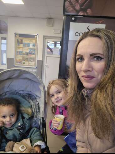 Eine Frau mit langen Haaren lächelt in die Kamera, neben ihr stehen zwei kleine Kinder – eines im Kinderwagen, das andere hält einen gelben Becher in der Hand; die Szene spielt in einem Innenraum.
