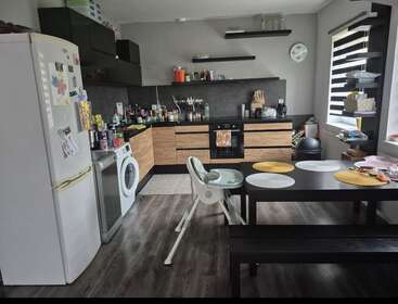 Dieses Bild zeigt eine moderne Küche mit Holzschränken, schwarzen Arbeitsplatten, einem Esstisch, einem Hochstuhl, einem Kühlschrank, einer Waschmaschine sowie verschiedenen Küchengeräten und -utensilien.