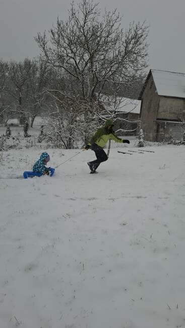 Una persona con chaqueta verde tira de un niño en trineo a través de la espesa nieve. Los árboles desnudos y los edificios cubiertos de nieve crean una escena invernal apacible.