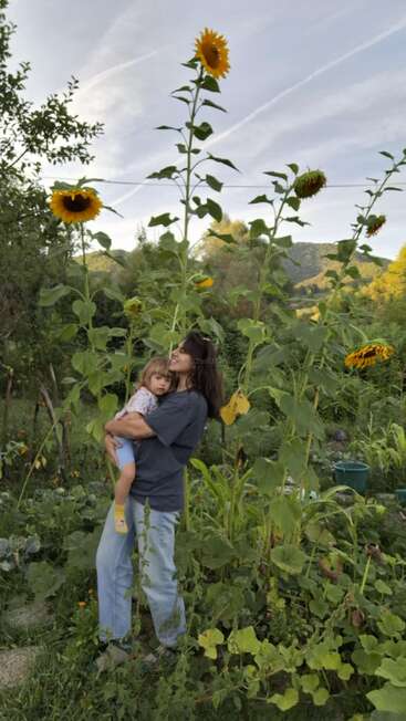 Una mujer sostiene a un niño pequeño entre altos girasoles en un exuberante jardín verde. De fondo, montañas, árboles y un tranquilo cielo azul.