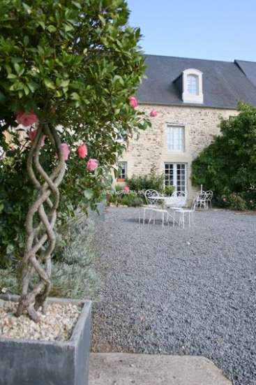 The image depicts a serene courtyard with a stone house in the background, featuring a gravel surface and a white table and chairs.