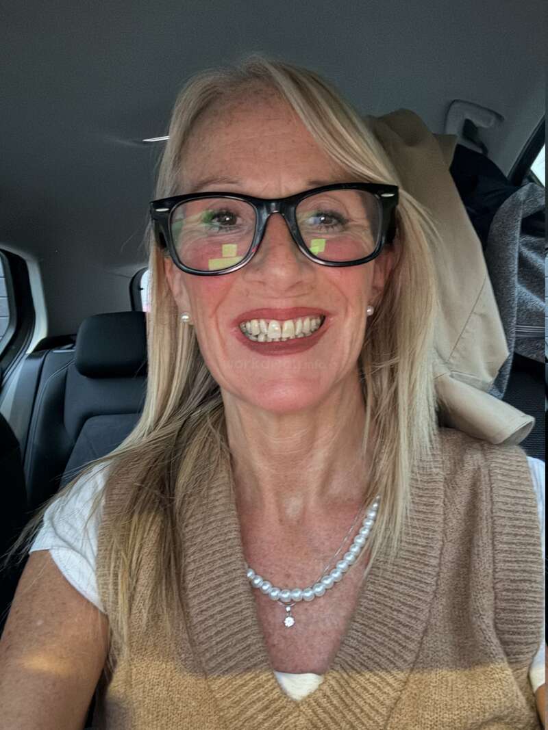 A smiling woman with blonde hair and glasses sits in a car. She wears a brown vest, white shirt, pearl necklace, and visible joy on her face.