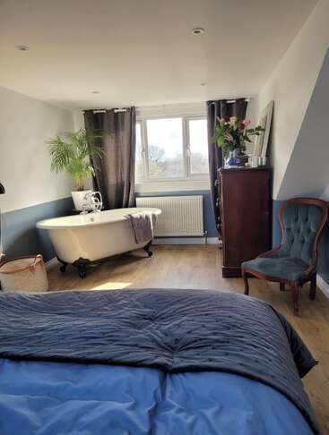 Quarto iluminado pelo sol com uma cama, banheira vintage, vaso de plantas, cadeira verde elegante, cômoda alta, cortinas escuras, piso de madeira e arranjo floral, criando um ambiente aconchegante.