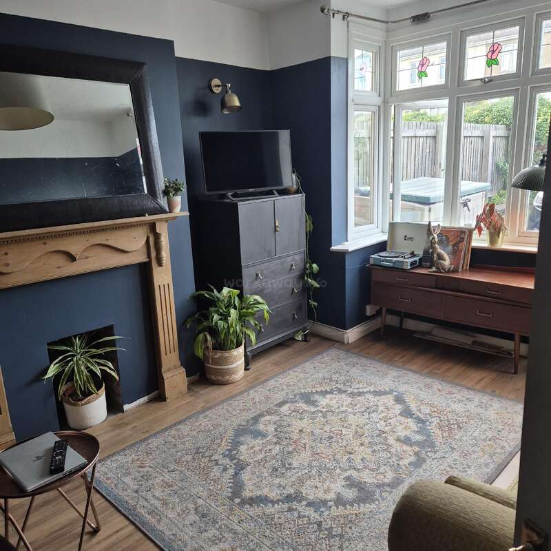 Uma sala de estar aconchegante com paredes azul-marinho, um grande espelho acima de uma lareira de madeira, plantas de interior, tapete estampado, gabinete de TV, mesa lateral e assentos na janela saliente.