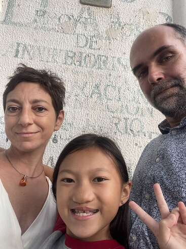 The image shows a family of three posing for a selfie in front of a white wall with green graffiti, smiling and making peace signs.