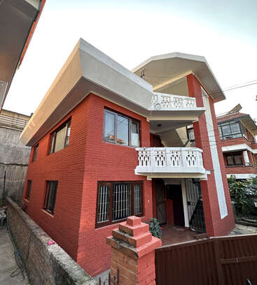Esta imagen muestra una casa de ladrillo rojo de dos plantas con barandillas blancas en los balcones, grandes ventanas, tejado en ángulo y una pequeña verja. En las inmediaciones se ven edificios vecinos.