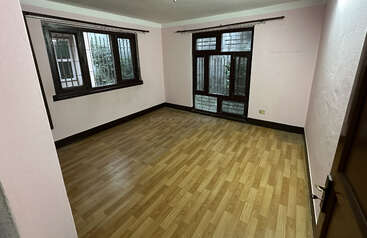 Una habitación vacía con paredes de color rosa claro, suelo de madera, grandes ventanas con marcos oscuros, barras de cortina y algo de luz natural que entra del exterior, creando un ambiente acogedor.