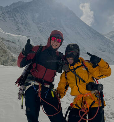 Dos alpinistas posan alegremente en una montaña nevada, ataviados con ropa brillante, cascos y cámaras, ante un dramático telón de fondo de imponentes picos nevados.