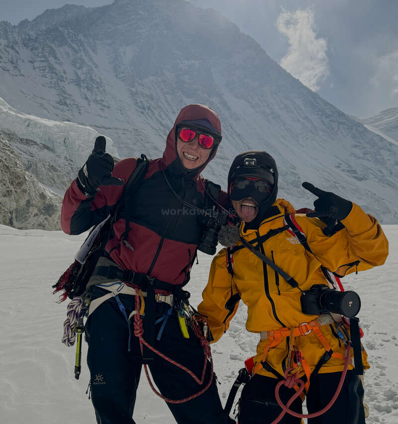 Dos alpinistas posan alegremente en una montaña nevada, ataviados con ropa brillante, cascos y cámaras, ante un dramático telón de fondo de imponentes picos nevados.