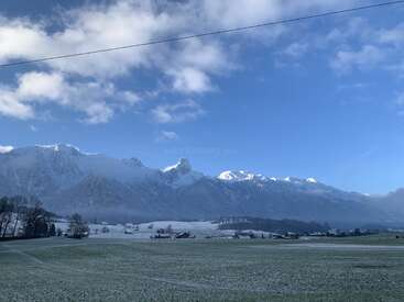 Las montañas nevadas se elevan majestuosas bajo un cielo azul brillante, mientras que un campo helado y casitas dispersas descansan apaciblemente en el tranquilo paisaje invernal de abajo.