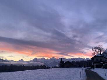Un impresionante amanecer de invierno pinta el cielo de tonos púrpura y naranja sobre casas cubiertas de nieve, graneros y majestuosos picos montañosos, creando una apacible escena rural.
