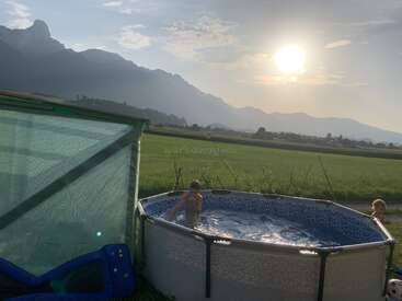 Dos niños juegan en una pequeña piscina redonda junto a un campo verde. Las montañas y un sol radiante crean una tranquila y pintoresca escena rural de fondo.